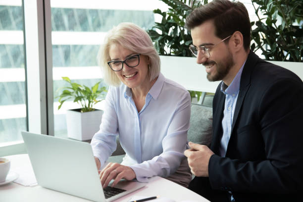 glücklicher leiter mittleren alters, der mit jungen kollegen am computer arbeitet. - trainee suit men business person stock-fotos und bilder