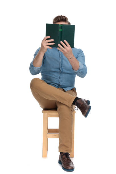 młody przypadkowy mężczyzna zakrywający twarz książką i czytaniem - sitting young men book isolated on white zdjęcia i obrazy z banku zdjęć