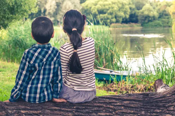 Photo of Two children, boy and girl, sitting on trunk of tree and watching at lake scenery