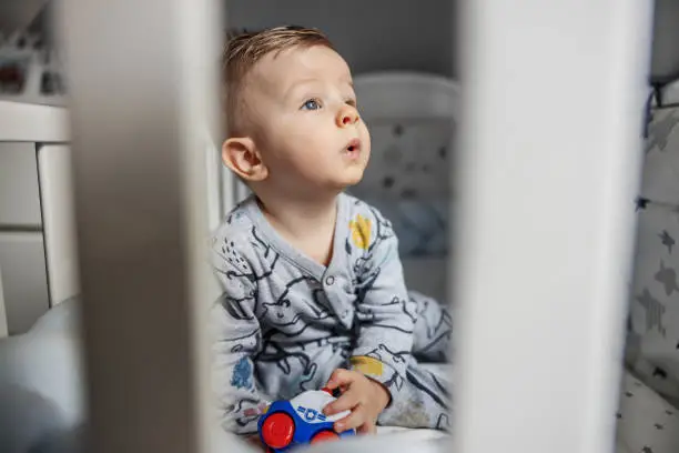 Photo of Puzzled charming blond baby boy sitting in his crib in the morning, holding his toy, looking at his parent and babbling.