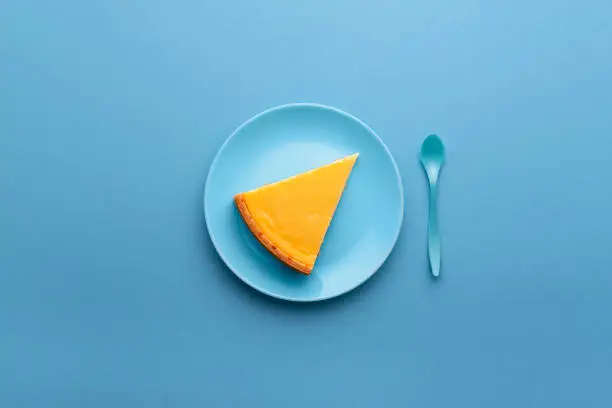 Single piece of cheesecake on a blue plate on blue background. Above view of dessert plate with cheesecake slice and blue spoon. Famous cheese cake.