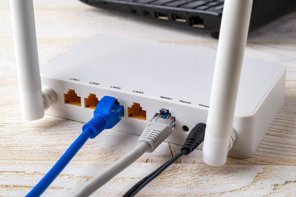 白い木製のテーブルのラップトップの近くの白いwi-fiワイヤレスルータに接続されたネットワークケーブルのクローズアップ。インターネット ケーブルが家庭やオフィスのテーブルに接続さ� - network connection plug network server computer cable wireless technology ストックフォトと画像