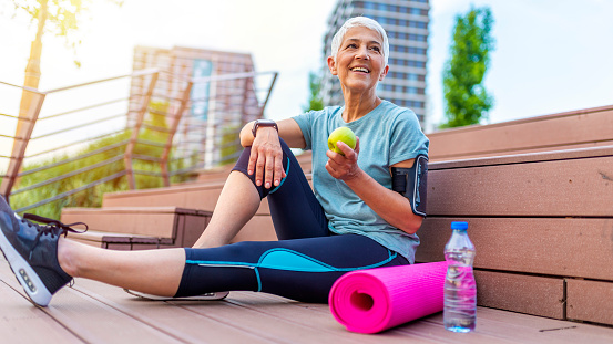 Sport mature woman sitting and resting after workout or exercise and eating apple on floor. Relax concept. Strength training and Body build up theme. Beautiful sporty woman eating apple