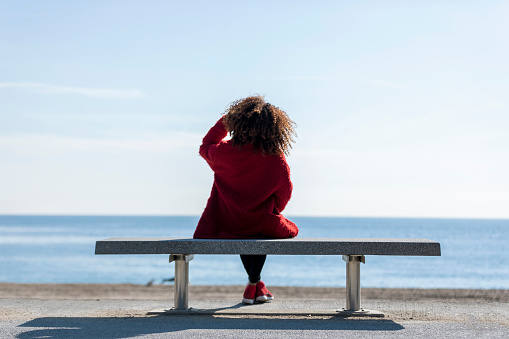 Rear view of a young curly woman wearing red denim jacket sitting on a bench while looking away to horizon over sea