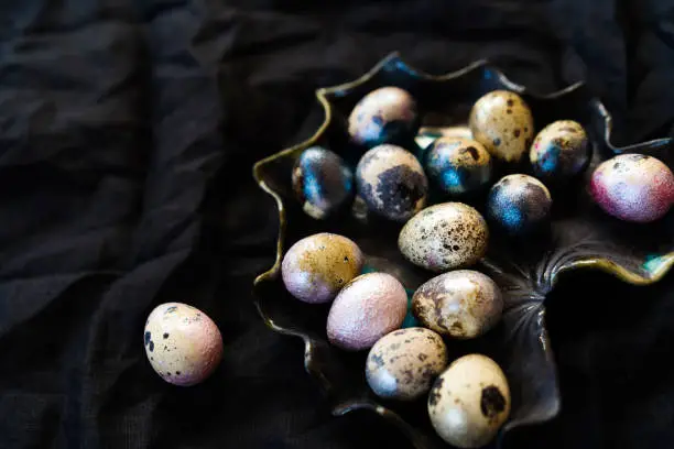 Colored painted pearl chicken and quail eggs of pink, silver, golden and blue color on black handmade ceramic plate background. Creative easter festive flat lay. Copyspace for text. Low key photo
