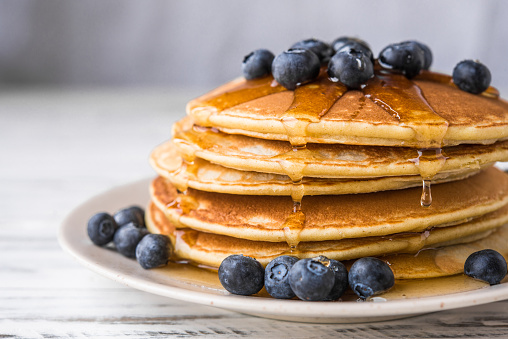 Close up of fluffy pancakes with maple syrup and blueberries against white wooden background
