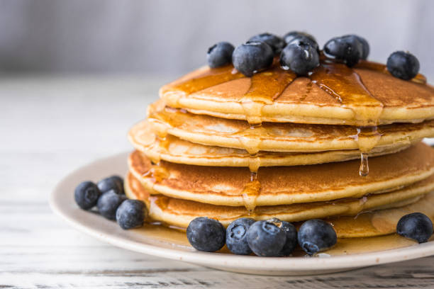 Close up of fluffy pancakes with maple syrup and blueberries against white wooden background Close up of fluffy pancakes with maple syrup and blueberries against white wooden background pancake stock pictures, royalty-free photos & images
