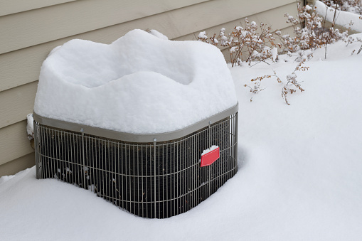 This image shows a close-up abstract view of an exterior air conditioner system unit, covered with fresh deep snow following a winter blizzard.