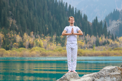 A young man in white practices yoga in the mountains. Pose Samasthiti namaskar