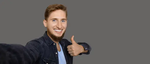 A bearded caucasian man with blue eyes who is making a selfie and gesturing the approbation sign while smiling on a grey background with freespace