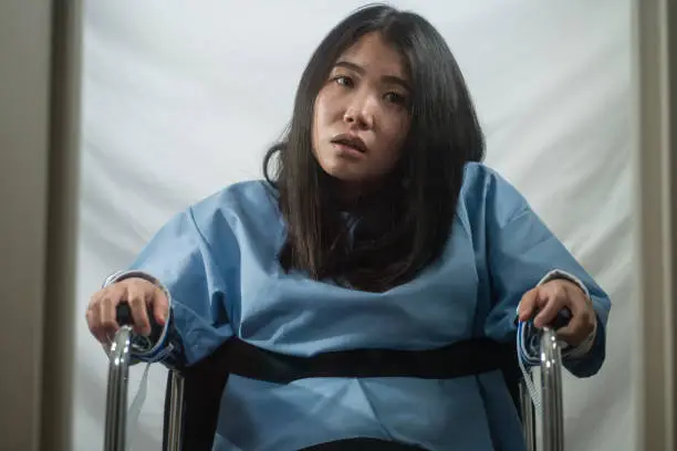 insanity asylum horror movie style portrait of young sick and psychotic Asian Chinese woman isolated and locked in mental hospital security cell sitting on wheelchair suffering schizophrenia