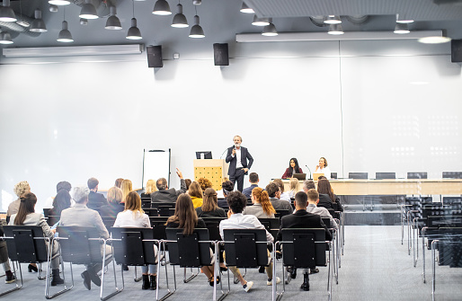 Mid adult businesswoman talking to group of seminar attendees in conference hall.