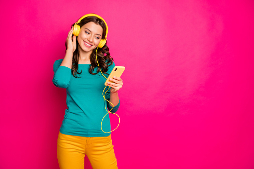 Photo of cheerful nice pretty cute charming girlfriend listening to music in her headphones, holding telephone with hands isolated pink vibrant color background