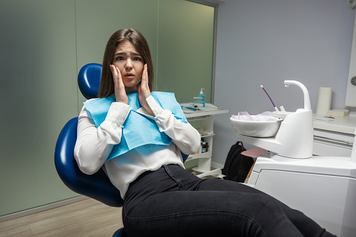 beautiful young scared patient woman sitting in dental chair during examination at dental office desperately touching her face looks frightened , healthcare concept.