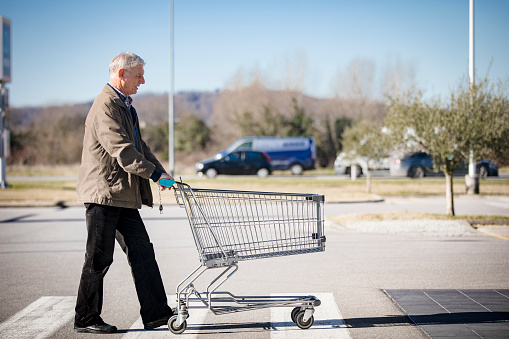 Senior Adult Man Pushing Empty Shopping Cart on Supermarket's Parking Lot.