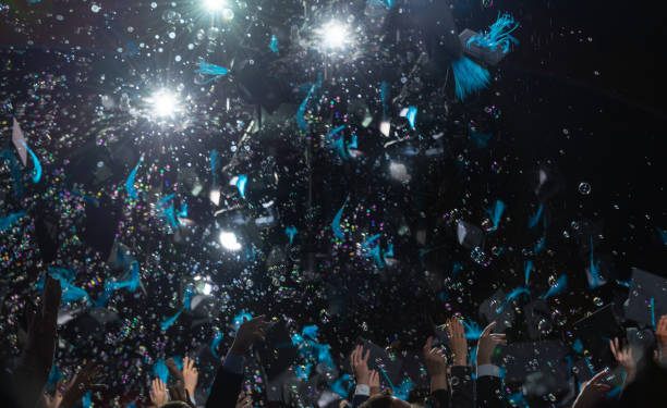Students throwing graduate caps during indoor graduation ceremony at university. Spotlights and bubbles on black background