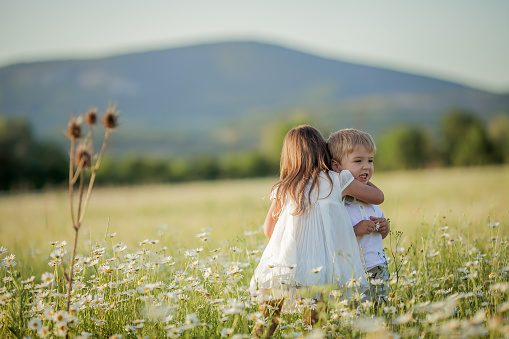 Cute beautiful young woman holding hands and play with little child girl walk in chamomile field with blooming flowers over nature background. Family lifestyle concept. Mom and baby daughter in grass