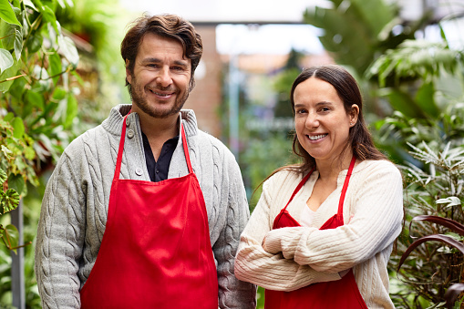 Portrait of smiling male and female owners in garden center. Confident woman with arms crossed standing by coworker. They are wearing red aprons.