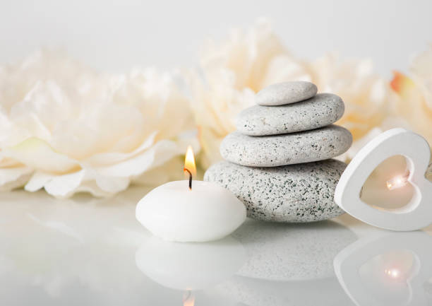 Stacked zen stones with white burning candle and blurred soft floral background, white glowing wooden heart light. Relaxing lifestyle concept.