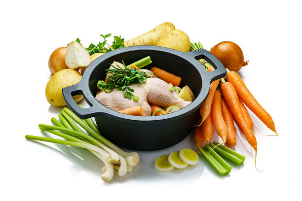 High angle view of a cooking pan filled with raw chicken bouillon ingredients isolated on white background. Ingredients for preparing chicken broth like raw chicken, carrots, kale, potatoes, celery and onions are also all around the pan. High resolution 42Mp studio digital capture taken with SONY A7rII and Zeiss Batis 40mm F2.0 CF lens