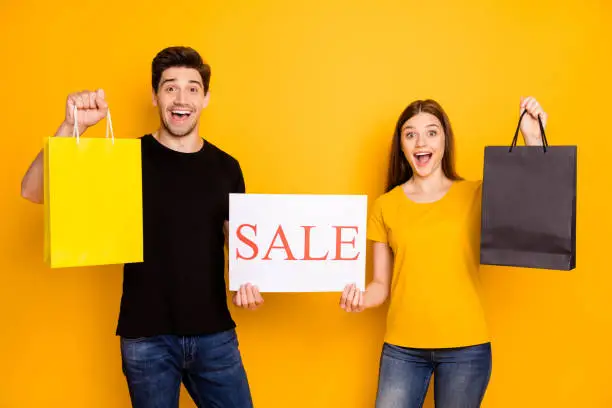 Photo of Photo of cheerful cute nice pretty sweet promoters showing you sheet of paper word sales written on holding packages discounted goods in black t-shirt jeans denim isolated vivid color background