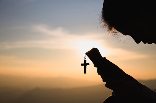 Siluette of Christian woman  holding a bible and wooden Christian cross necklace while praying to God.