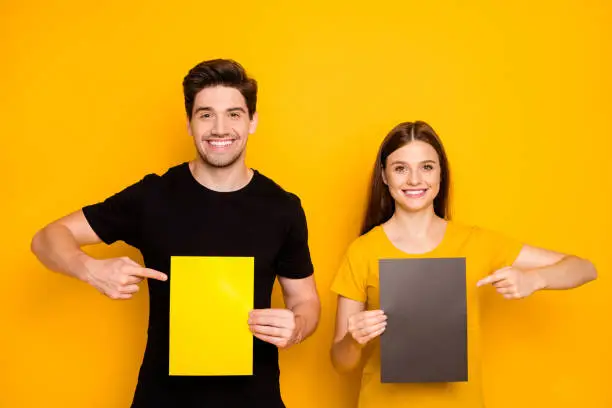 Photo of Photo of cheerful positive cute pretty sweet gorgeous two people working as advertisers together pointing at empty sheets of paper with forefingers isolated over vibrant color yellow background