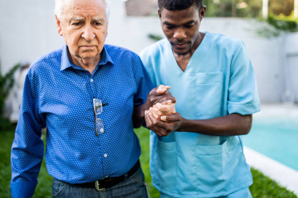 Black male nurse helping senior man to walk outdoors Black male nurse holding senior's man hand and helping him to walk in backyard of nursing home walking aide stock pictures, royalty-free photos & images