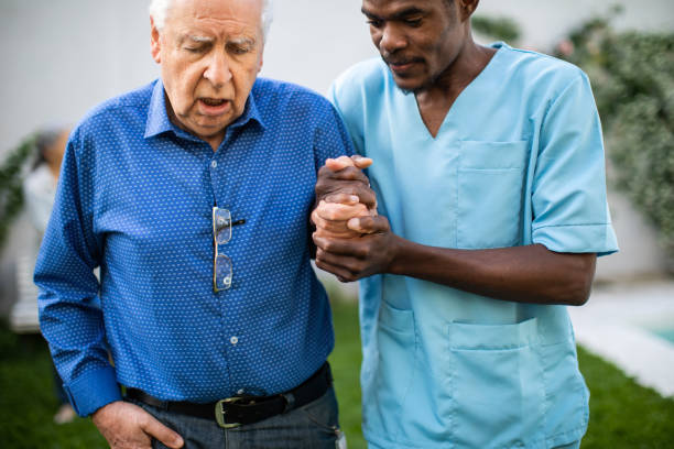 Black male nurse assisting senior man to walk outdoors Black male nurse holding senior's man hand and helping him to walk in backyard of nursing home walking aide stock pictures, royalty-free photos & images