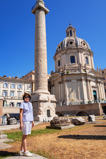 Mature woman standing in front of Trajan's Column on the acient Rome archaeology site. The Chiesa del Santissimo Nome di Maria al Foro Traiano Catholic church in the background