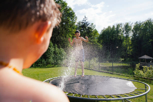 Happy beautiful Caucasian teenage young boy is jumping on a trampoline near a forest while his brother is spraying him with a sprinkler on a sunny hot day on a lawn in the backyard stock photo
