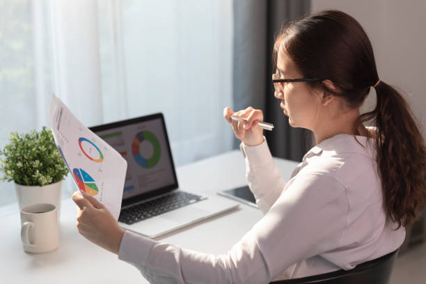 istock Young employee in eyeglasses working at office on laptop while sitting at table, Business woman analyze document in her hands, Graphs and diagram on notebook screen. 1207059442