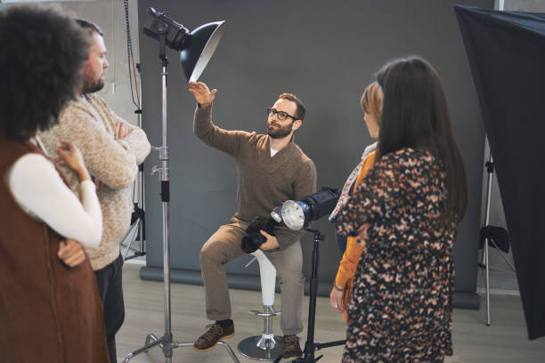 istock Young attractive bearded tutor sitting on chair in studio, holding camera and explaining something about photography while attendees standing and looking at him. 1207071990