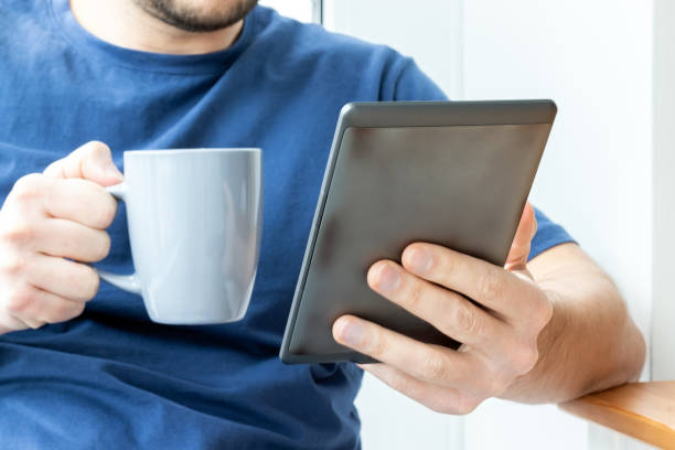 мужчина в синей футболке держит в одной руке серую электронную книгу электронного читателя, а в другой – чашку. чтение книги или новостей н� - e reader digital tablet cafe reading стоковые фото и изображения