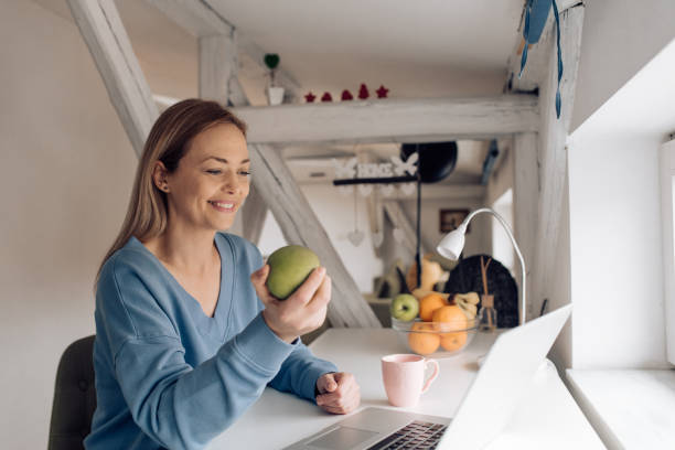 istock Beautiful woman eating apple and using laptop at home 1207073608