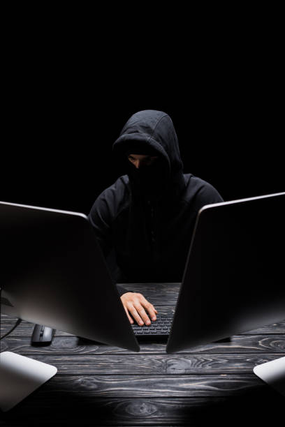 istock hooded man in mask sitting near computer monitors isolated on black 1207075773