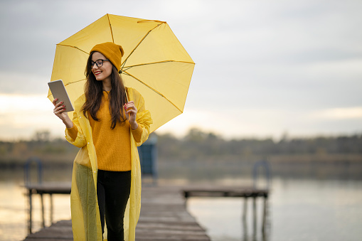 Happy young woman holding yellow umbrella, wearing yellow raincoat, walking on lake pier and working on digital tablet