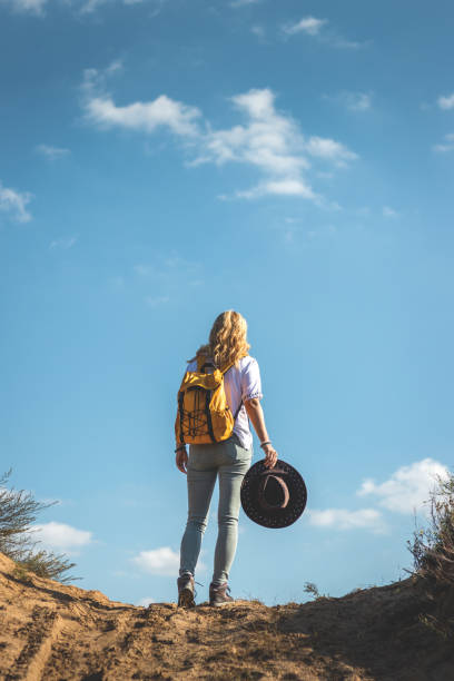 Woman with backpack and hat standing outdoors against blue sky