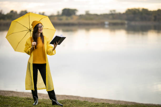 Female student with yellow umbrella wearing yellow raincoat and reading a book near lake