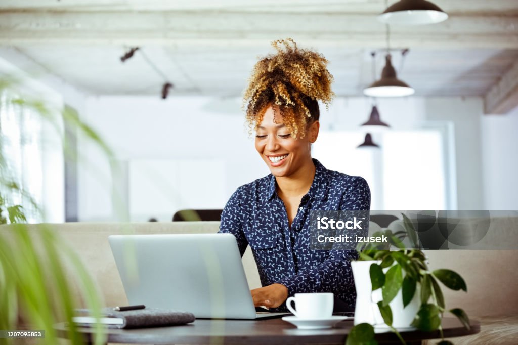 Smiling businesswoman using laptop in office Smiling mid adult businesswoman using laptop in office. Skilled corporate worker is having curly hair. She is working on investment plans. Office Stock Photo