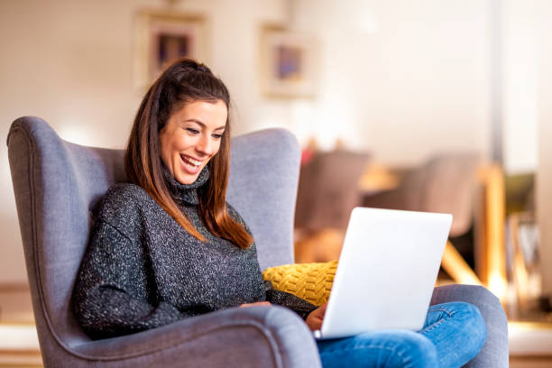 Happ young woman using notebook while working from home Shot of smiling woman sitting in the armchair and using laptop while working from home. Home office. happ stock pictures, royalty-free photos & images