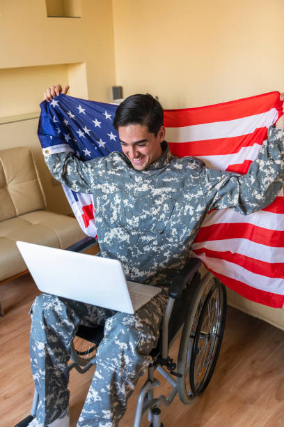istock Retired Soldier With Physical Disabilities is Using a Laptop and Holding US Flag 1207112469