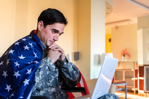 istock Retired Soldier With Physical Disabilities is Using a Laptop and Holding US Flag 1207112673