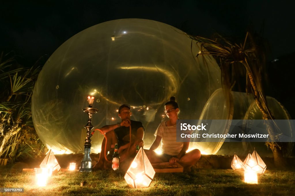 Badung - A couple sitting in front of a bubble hotel A couple sitting outside of a transparent bubble hotel, surrounded by candle light and smoking shisha, next to Nyang Nyang Beach, Bali, Indonesia. Luxury holiday. Romantic getaway. They are enjoying Bubble Stock Photo