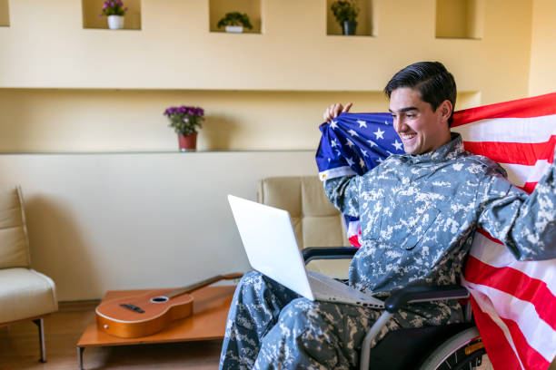 istock Retired Soldier With Physical Disabilities is Using a Laptop and Holding US Flag 1207112964