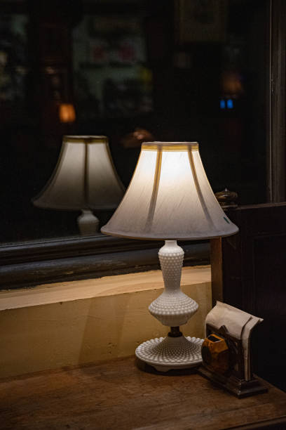 istock Elegant white antique table lamp closeup on dark blurry reflection. Lamp silhouette blurred reflection in glass window. Antique white lampshade glowing in darkness. 1207115142