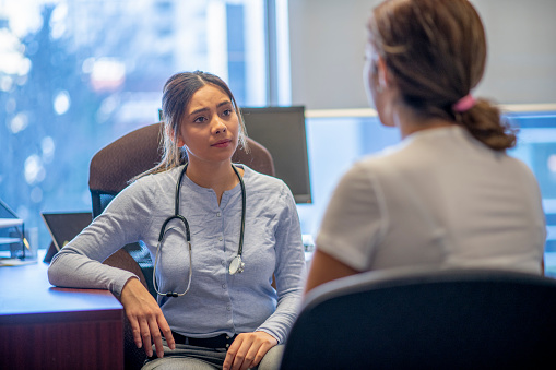 A female mixed race doctor, consults with a young female patient in her office.  She is seated at her desk, wearing a light blue semi-casual shirt, has a stethoscope around her neck and is leaning on her desk with her elbow.  She is listening attentively to the patient describe her symptoms.  The patient is dressed casually and has her back to the camera.