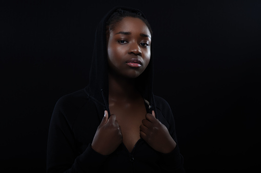 Confident woman with dark skin looking down. Cool attitude wearing hoodie. High quality low-key studio portrait.