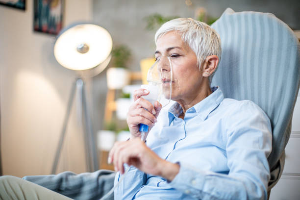 Senior woman doing inhalation. Portrait of a beautiful senior woman with grey hair doing inhalation at home. She is using inhaler for the treatment, inhaling through inhaler mask. medical oxygen stock pictures, royalty-free photos & images