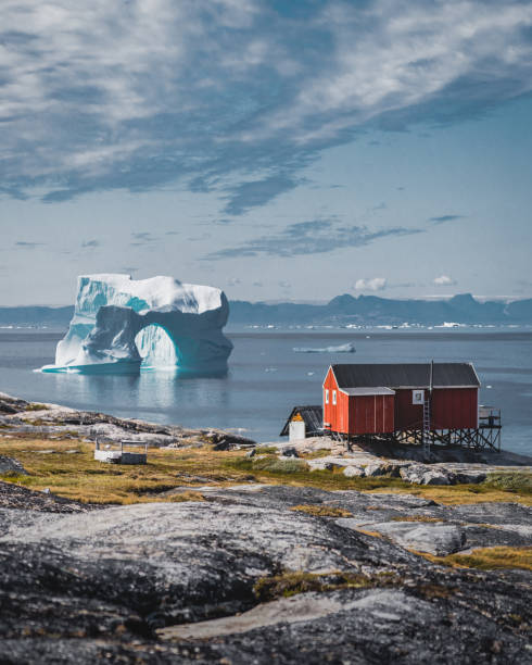 góra lodowa z łukiem w wodach grenlandii antarktyki na tle gór półwyspu arktycznego. małe kolorowe domy w słoneczny dzień z błękitnym niebem i chmurami. - greenland zdjęcia i obrazy z banku zdjęć